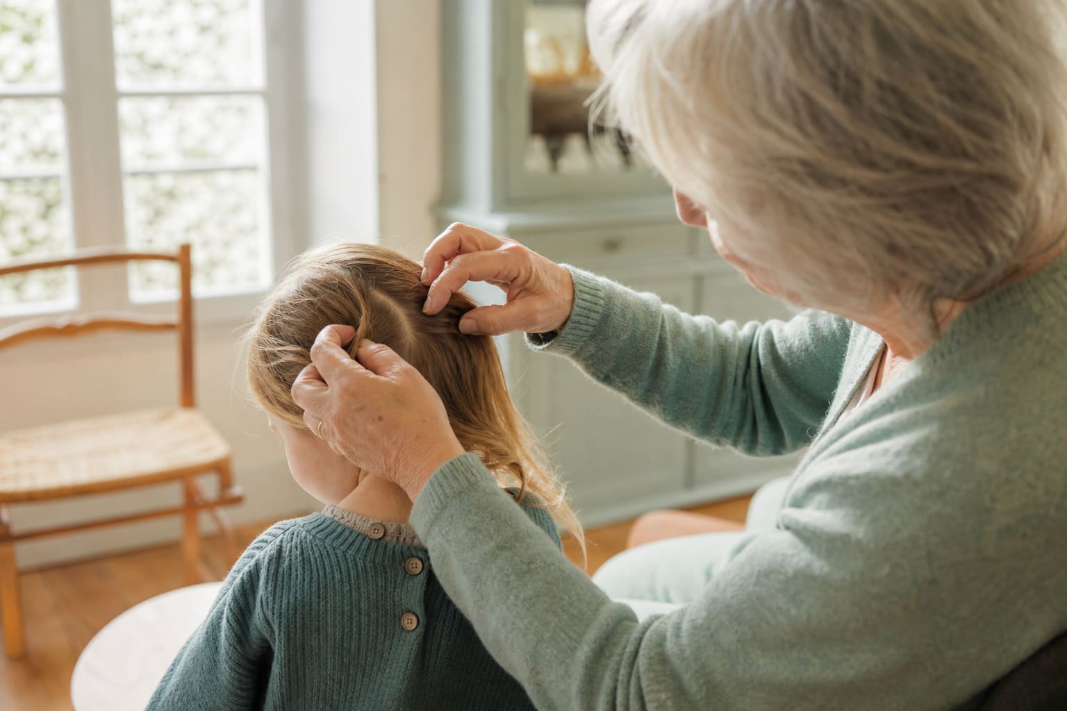 Grand-parent et enfant assis côte à côte près d’une fenêtre lumineuse, cheveux écartés pour un contrôle