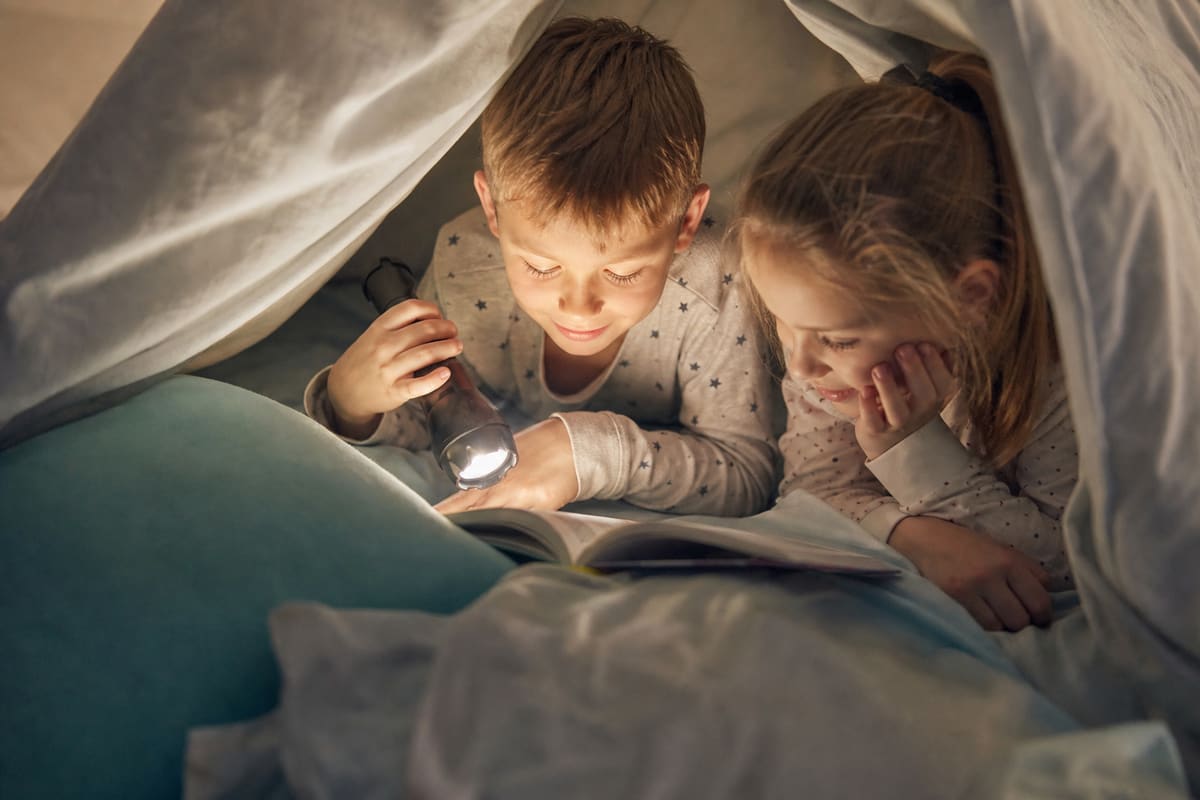 Deux enfants en pyjama allongés côte à côte sous une couverture, lisant un livre à la lampe torche