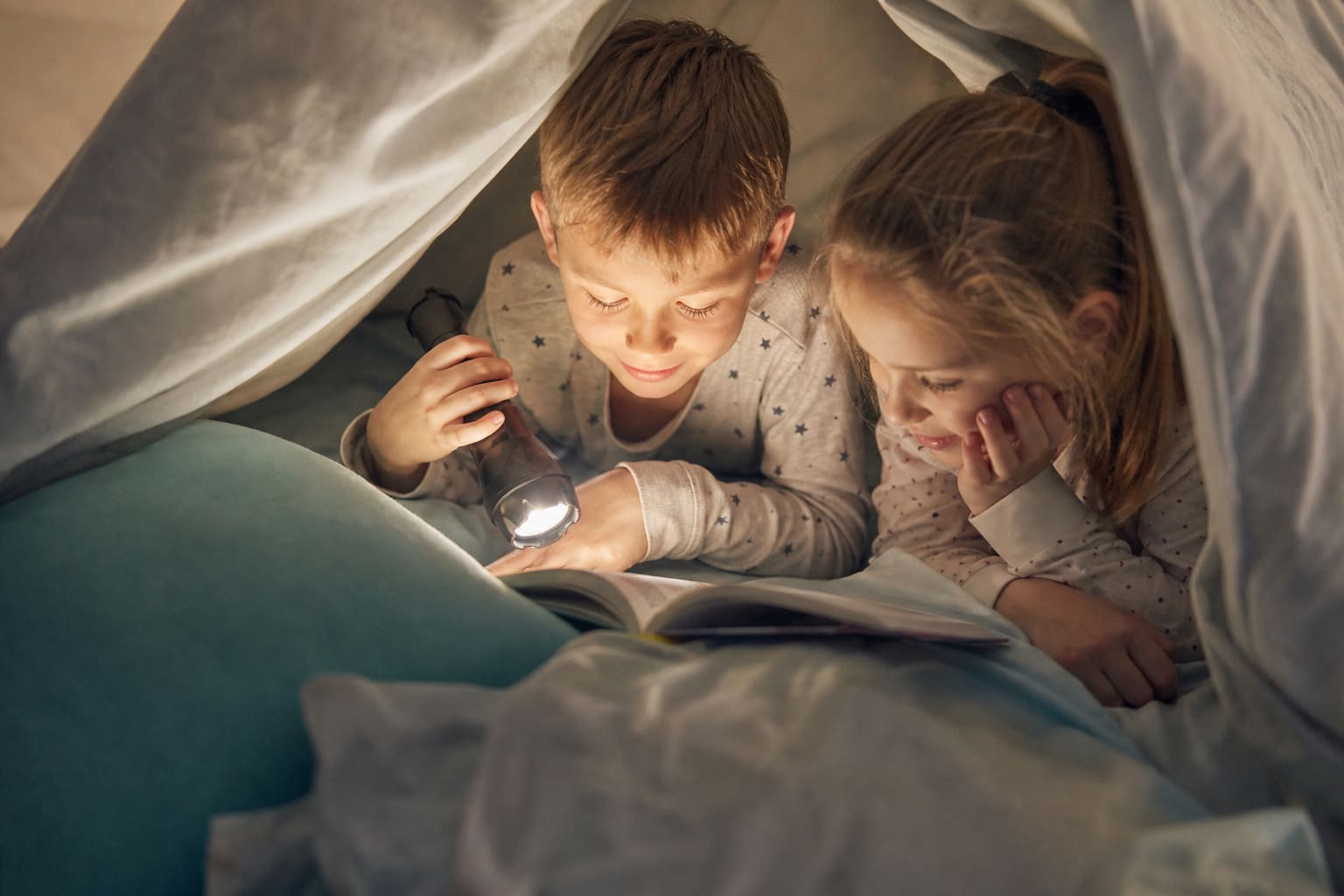Deux enfants en pyjama allongés côte à côte sous une couverture, lisant un livre à la lampe torche